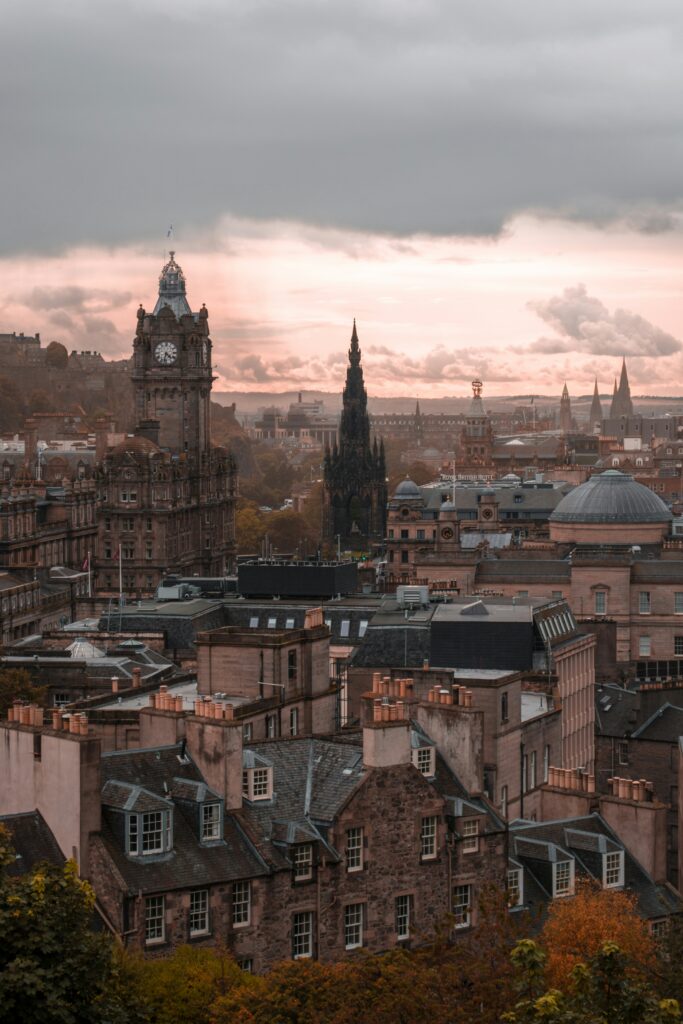 Overlooking view of Edinburgh, with some orange and green trees in autumn, and some sunlight peeking in through the clouds to shine some light on the city and clock tower.