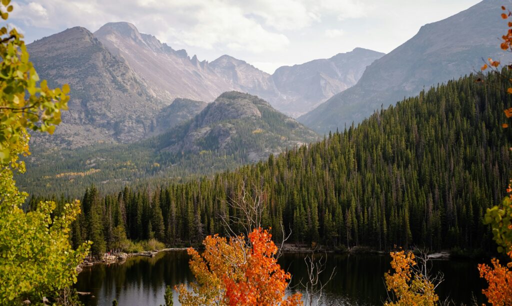 A view of mountains cascading throughout the image with tall pine trees and some crimson-colored trees in the foreground showing this photo was taken suring autumn.