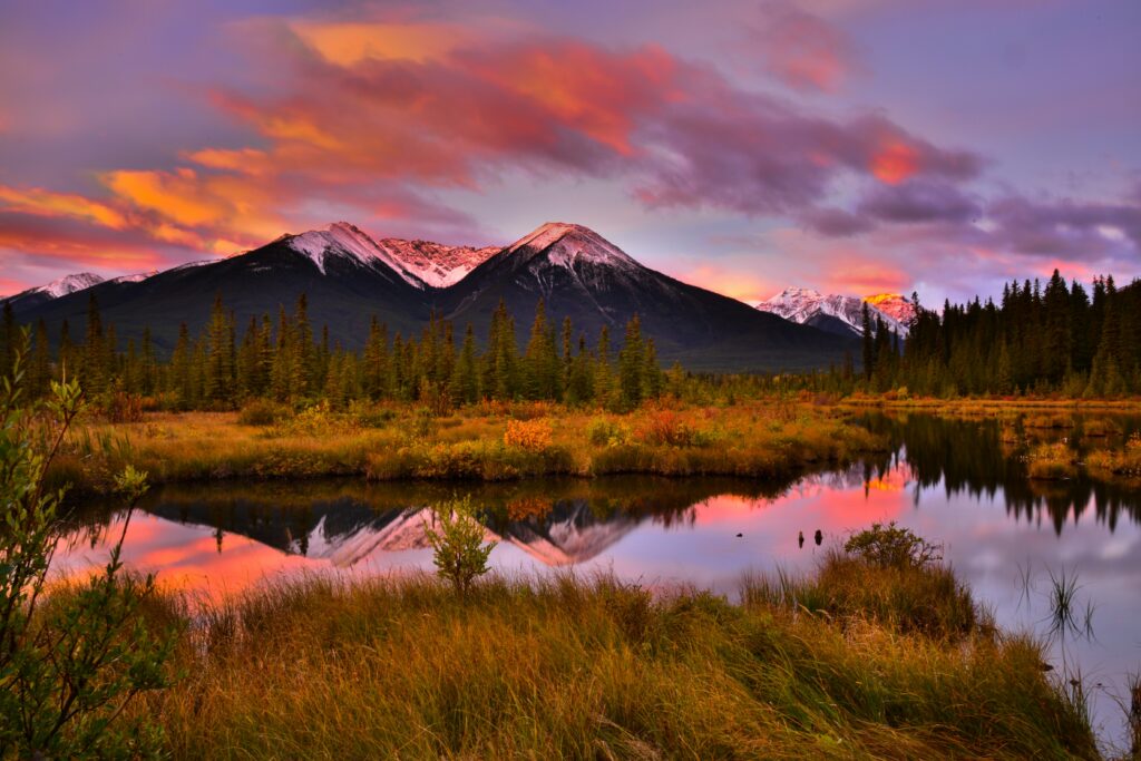 A scenic view of snow-peaked mountains during a sunset, creating a purple and orange sky atop the changing colors of the autumn trees while reflecting off a river.