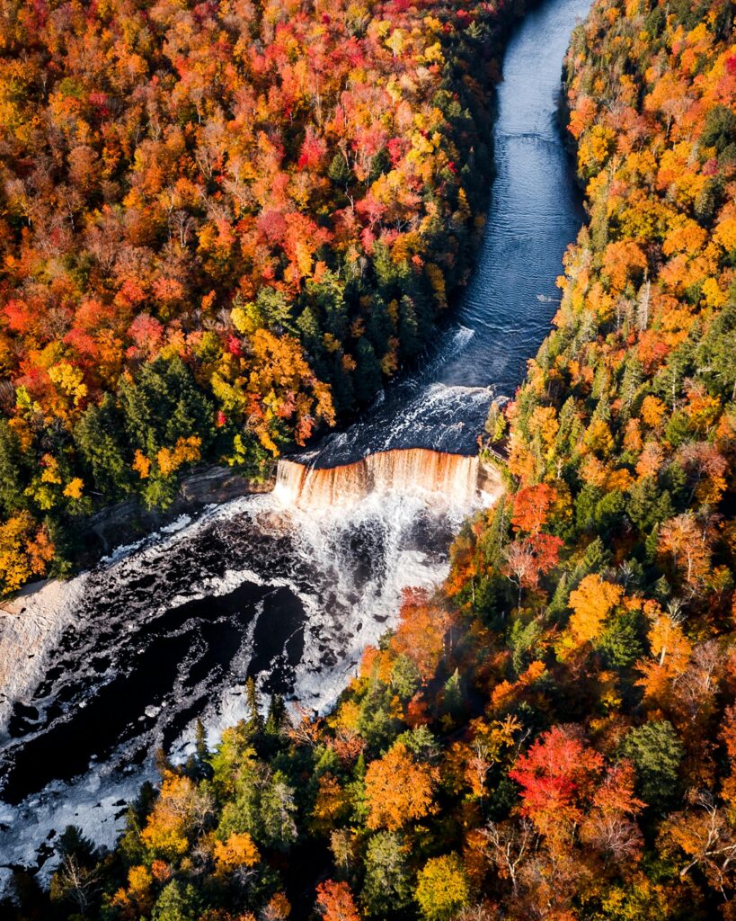 Birdseye view of a waterfall with a dark, navy, and silver blue river bookended with trees full of sutumn colors such as red, green, yellow, and orange.